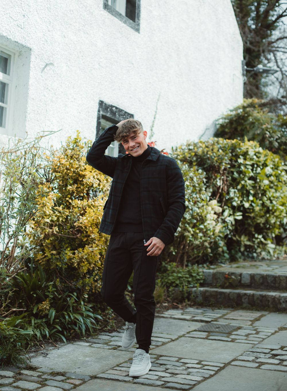 Casual street style pose in black outfit against white building with garden.