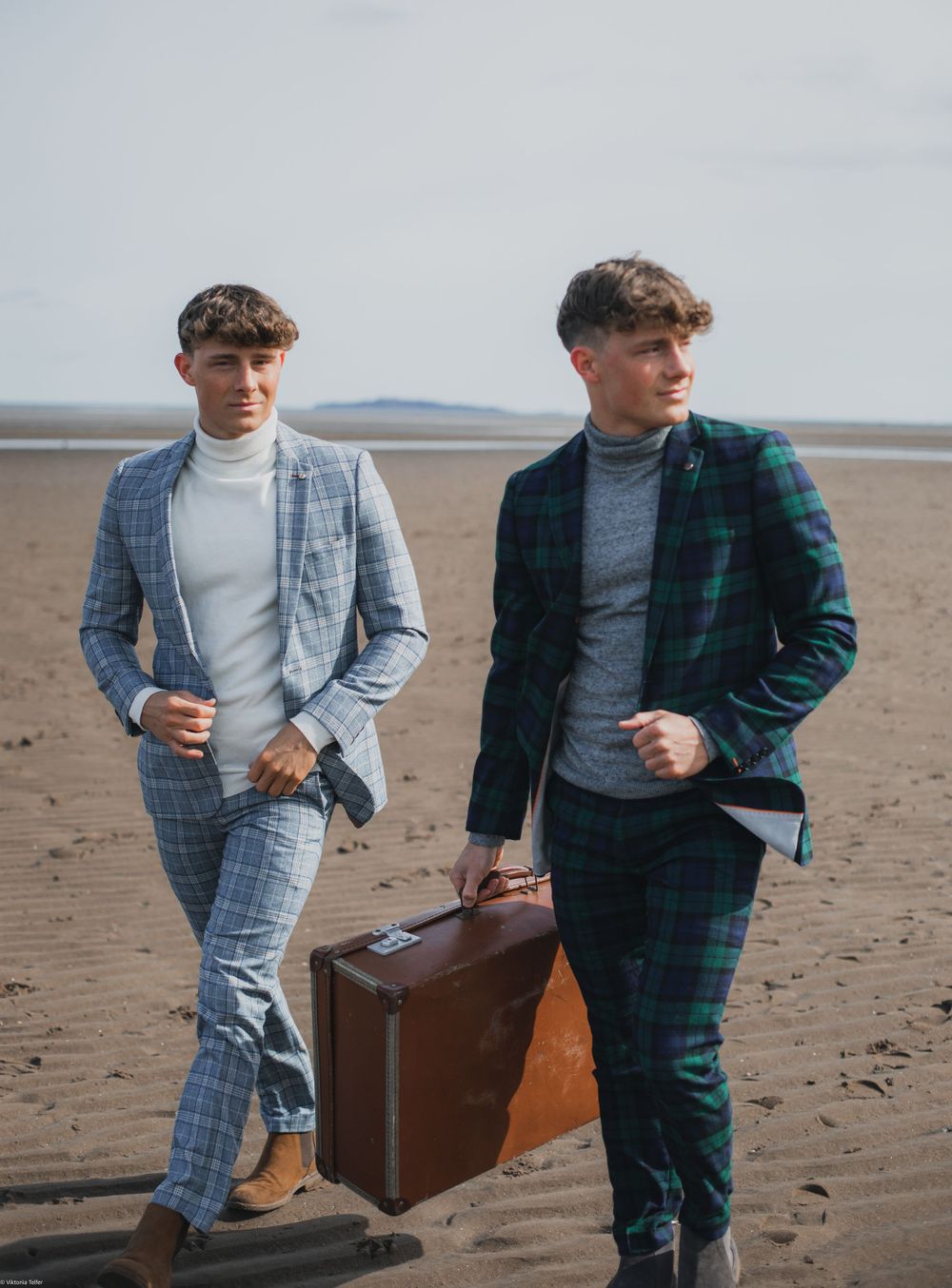 Two people in stylish suits walking on beach carrying vintage leather suitcase.