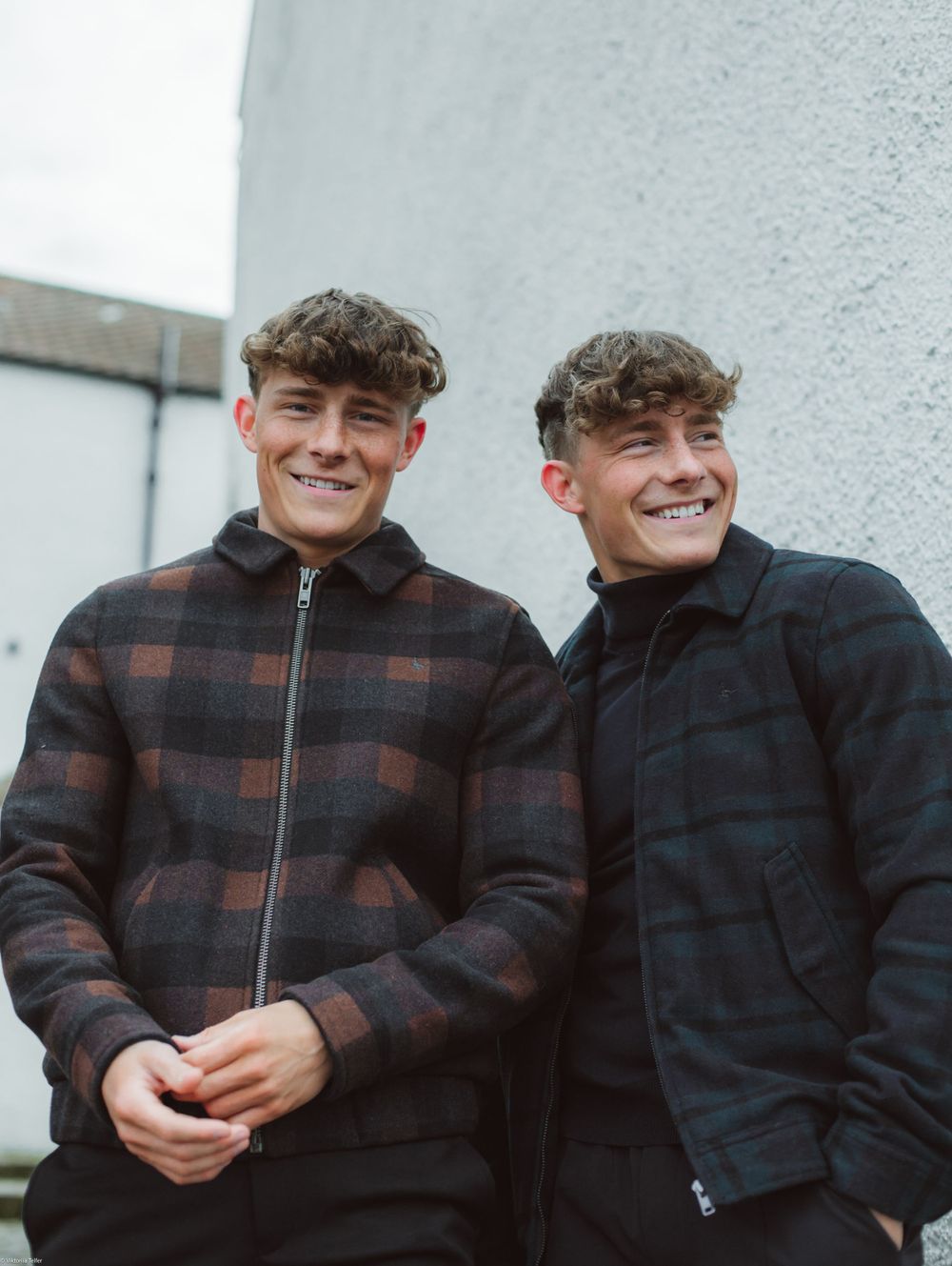 Two friends in plaid jackets smiling against white wall outdoors.