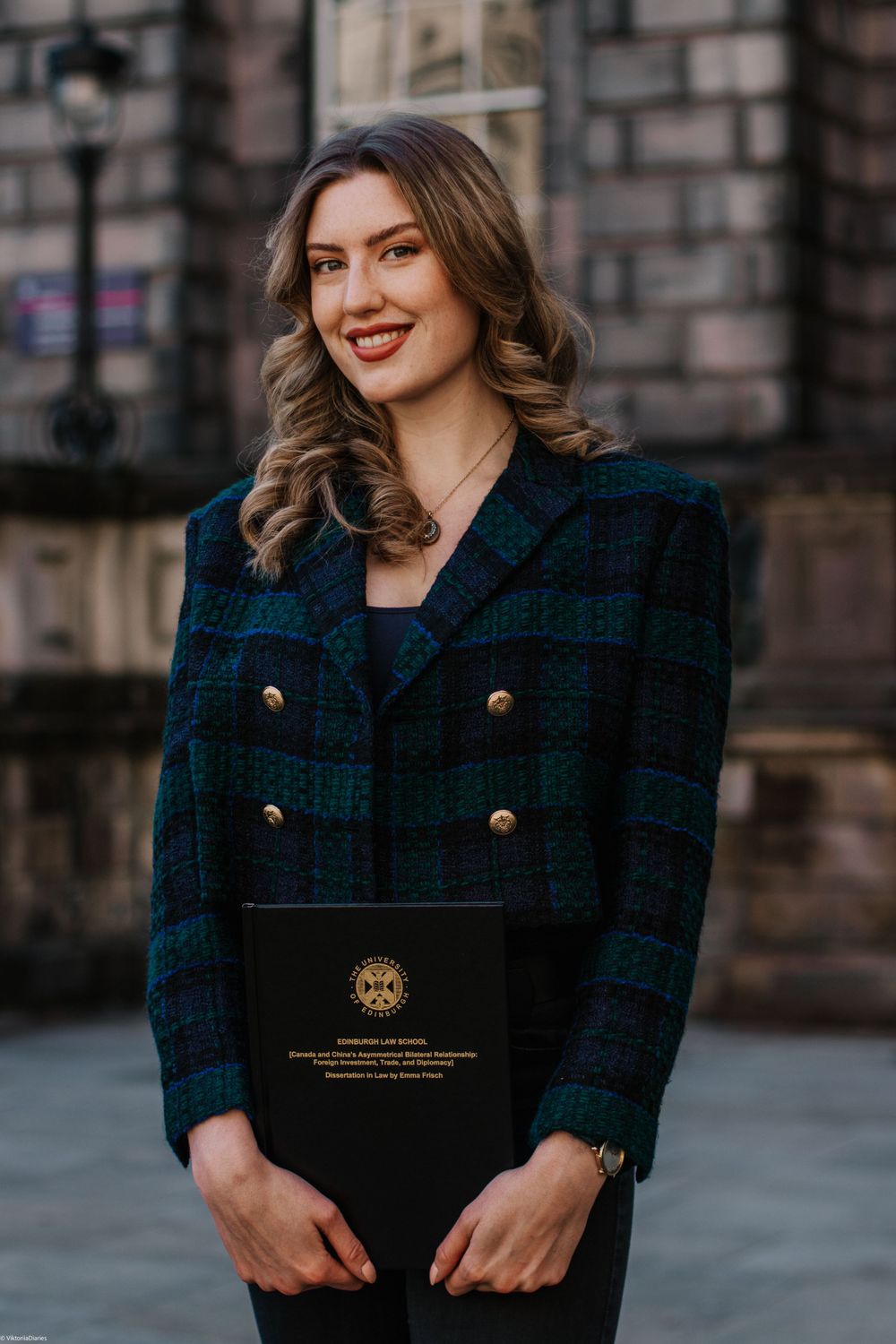 Graduate in plaid blazer holds diploma against stone building backdrop.