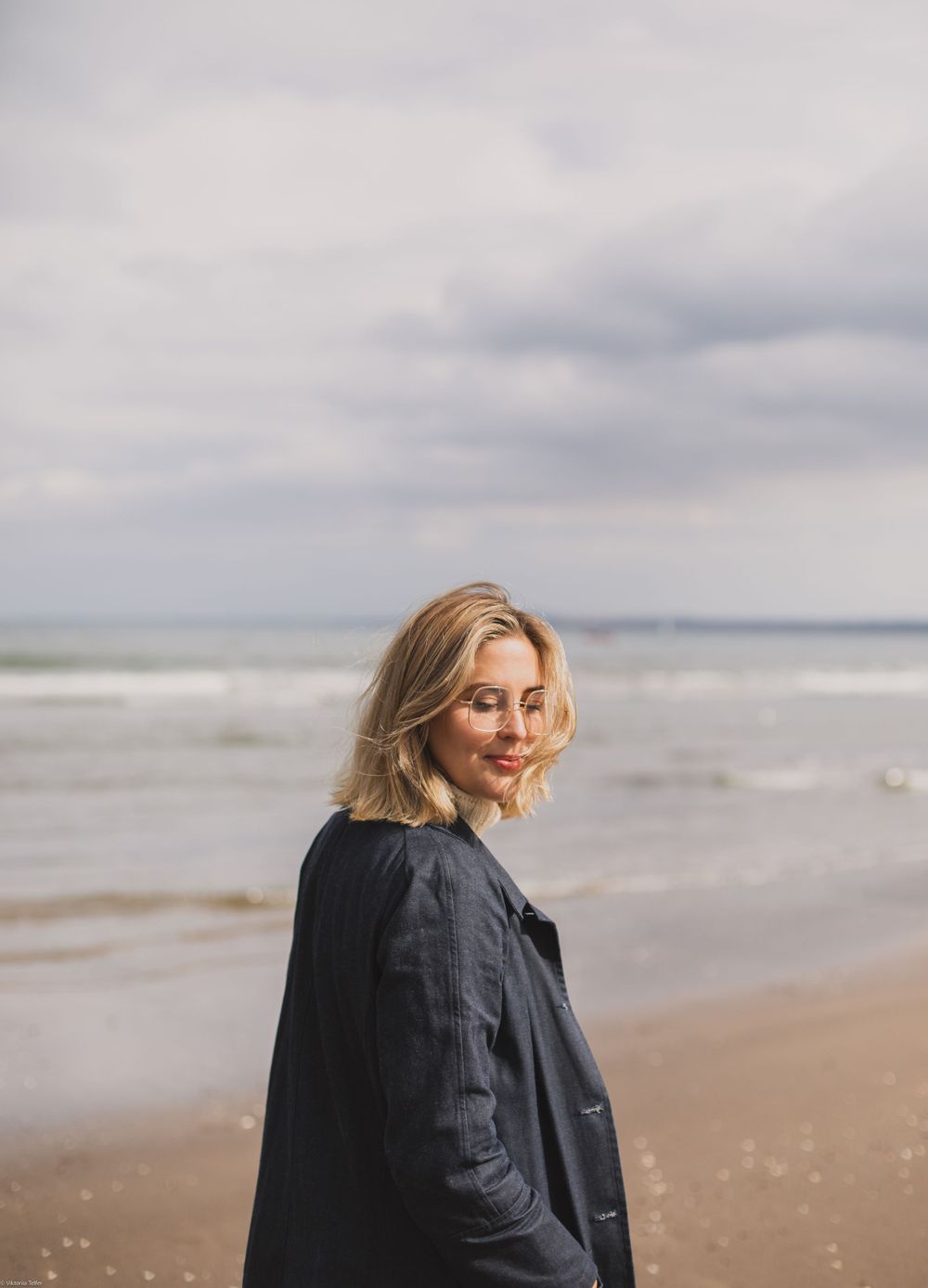 Woman in black coat stands on windswept beach during overcast day.