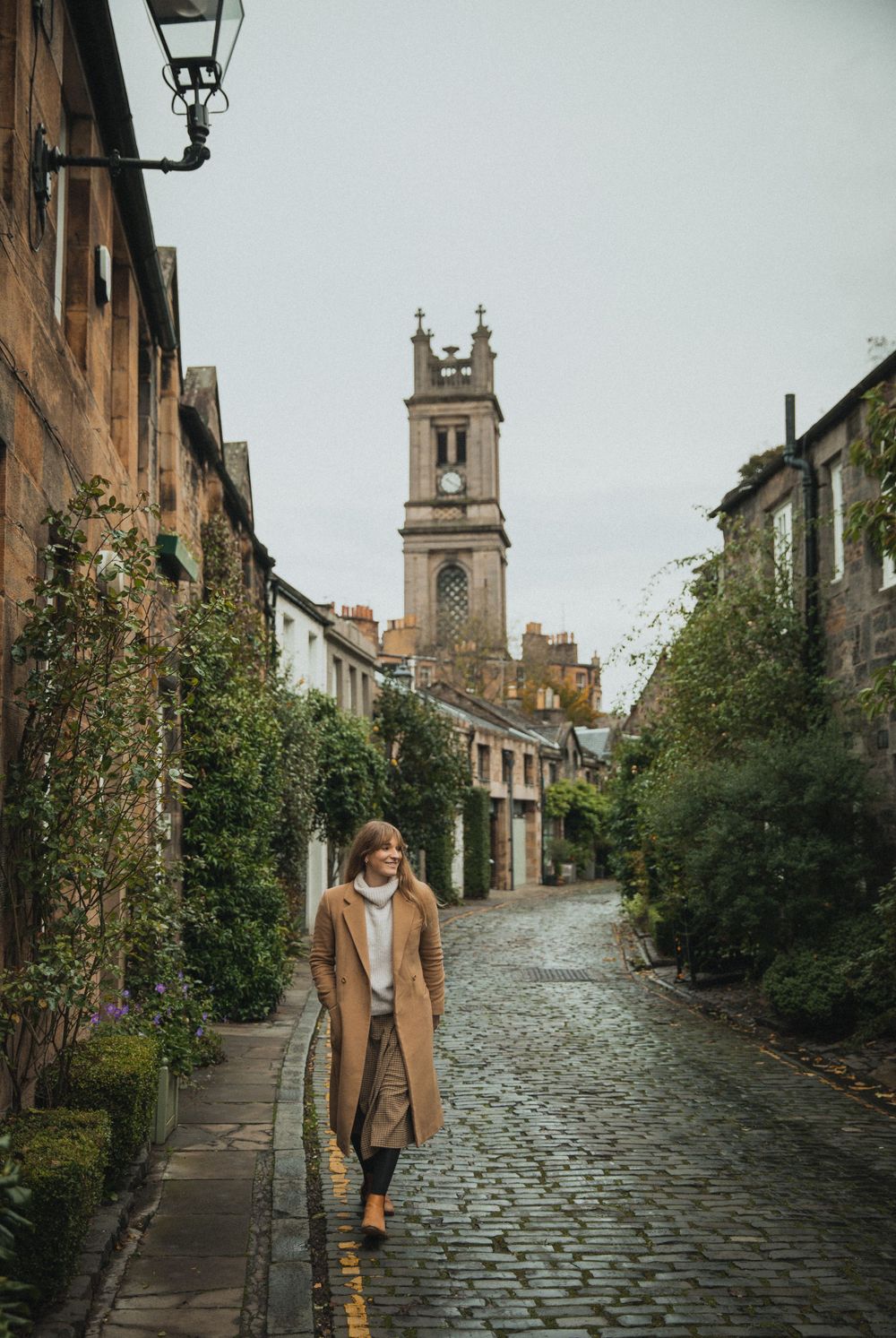 Person in camel coat walking on cobblestone street with historic church tower in background.