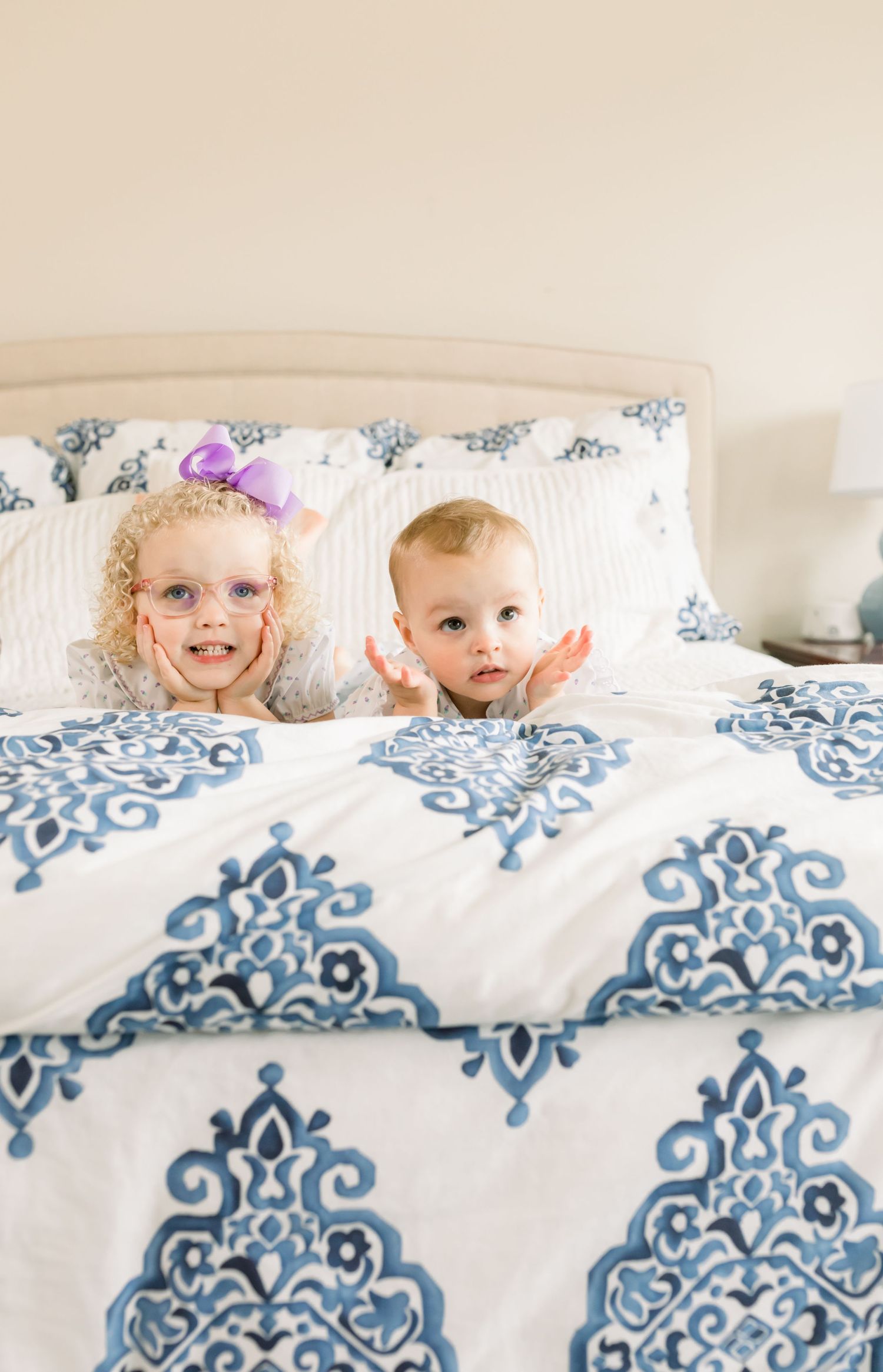 Two young children peeking out from behind white pillows on a patterned bed.