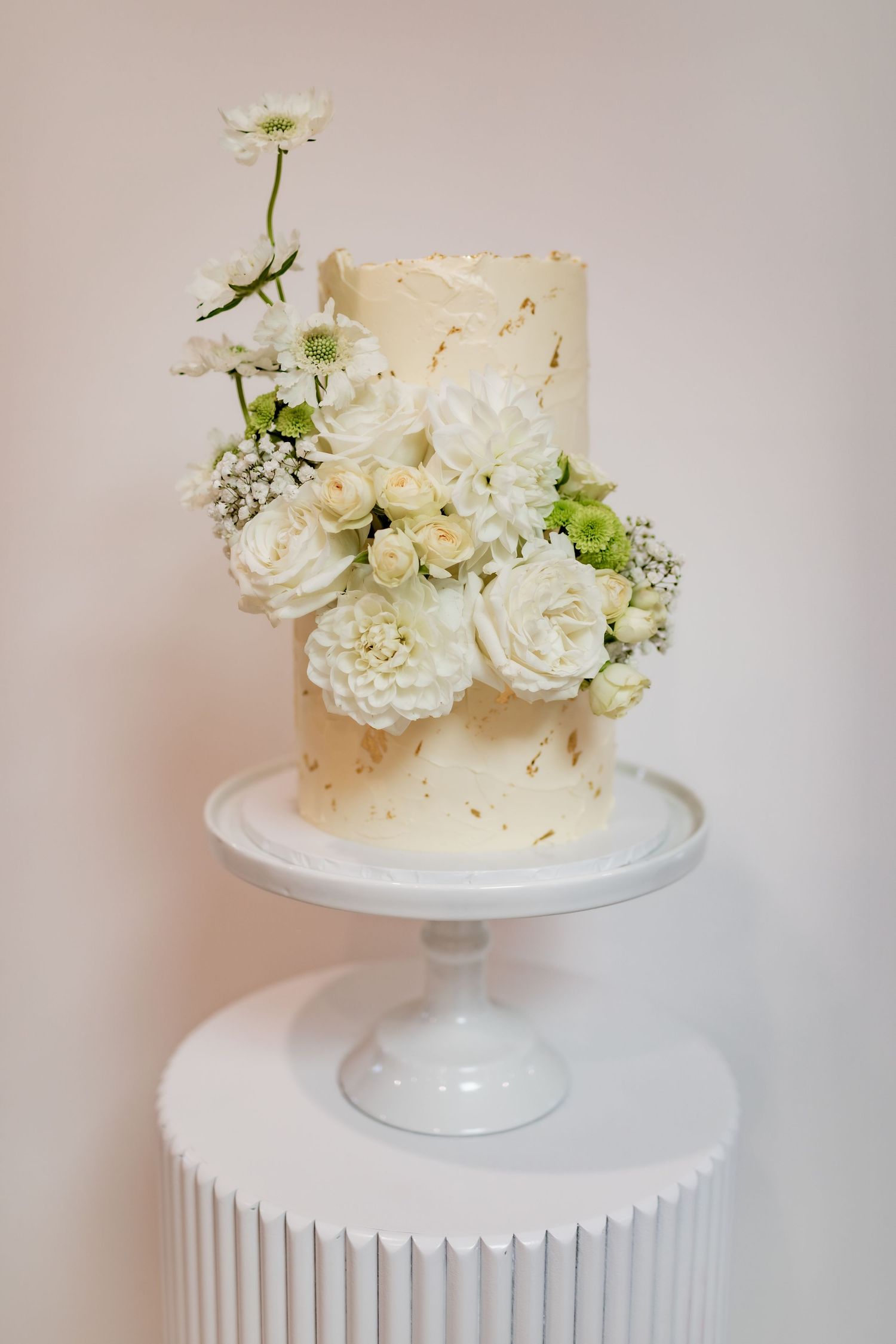 Single tier white wedding cake decorated with fresh flowers on a pedestal stand.