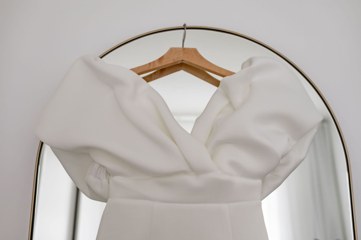 White wedding dress hanging on wooden hanger against arched mirror.