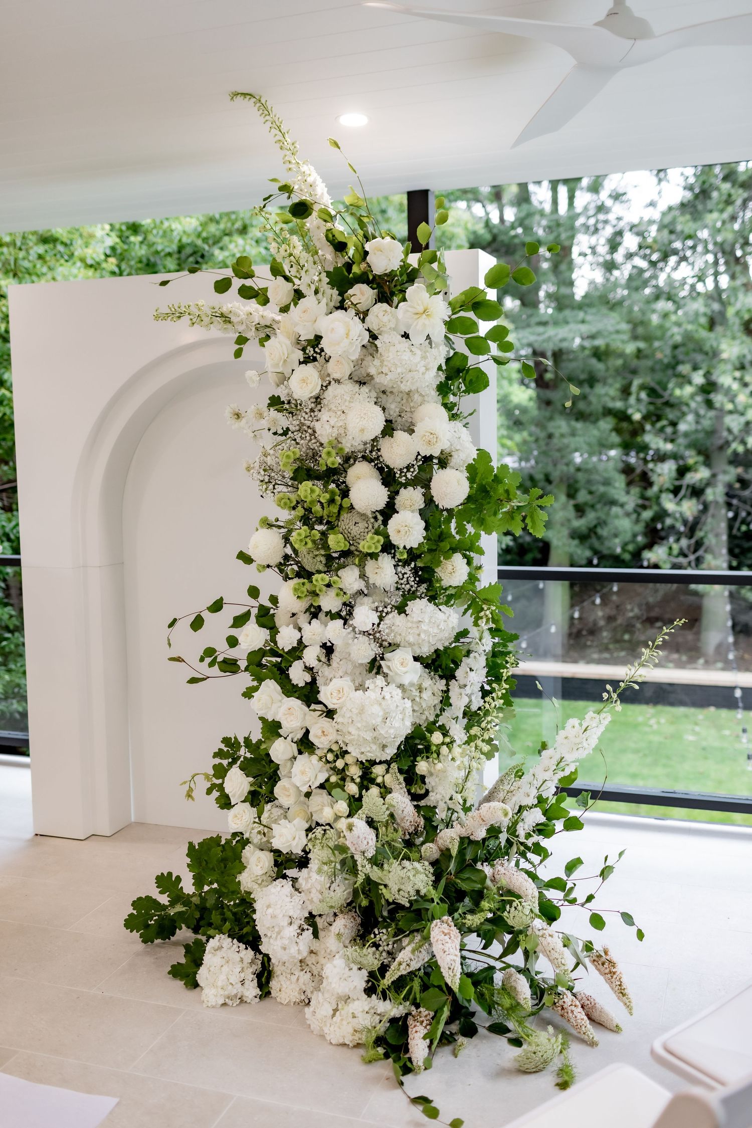 Elegant white floral arrangement with greenery cascading along a white arch backdrop at a wedding ceremony.