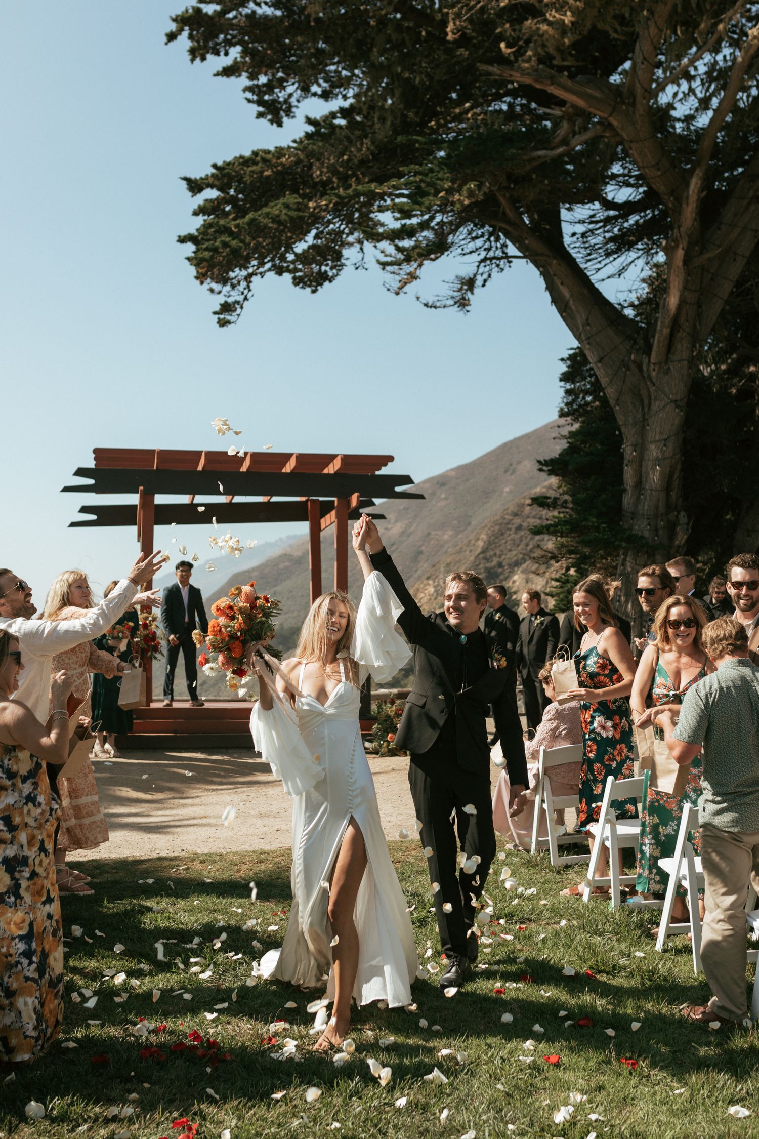 Wedding guests throw confetti and celebrate joyfully during a seaside ceremony under a wooden pergola.