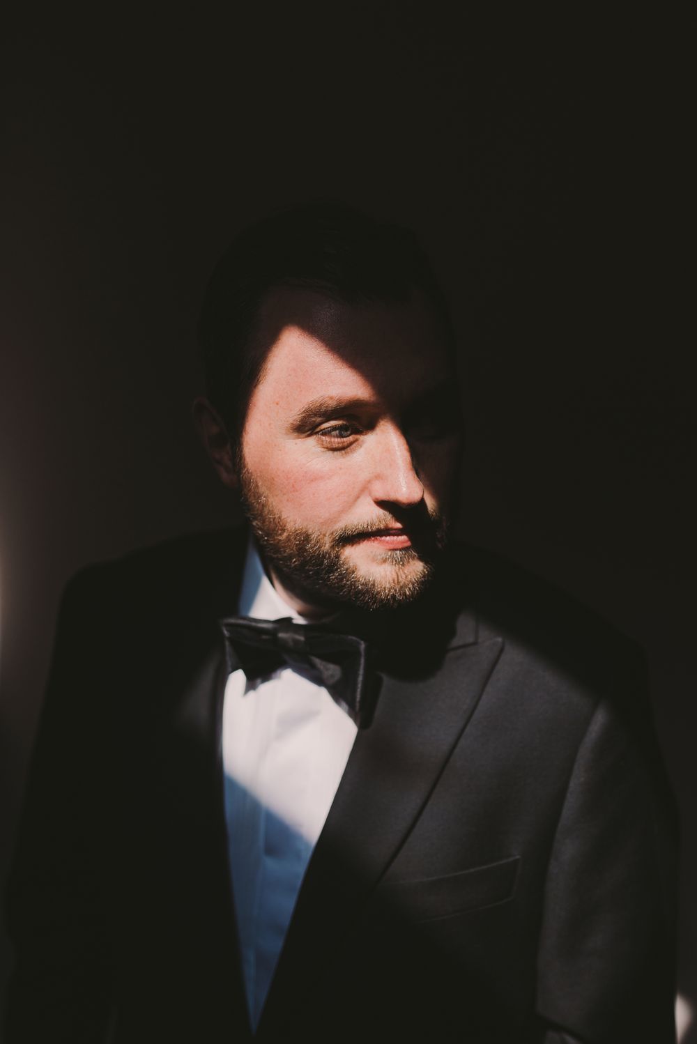 Dramatic close-up portrait in dark lighting showing partial face with beard in formal attire.