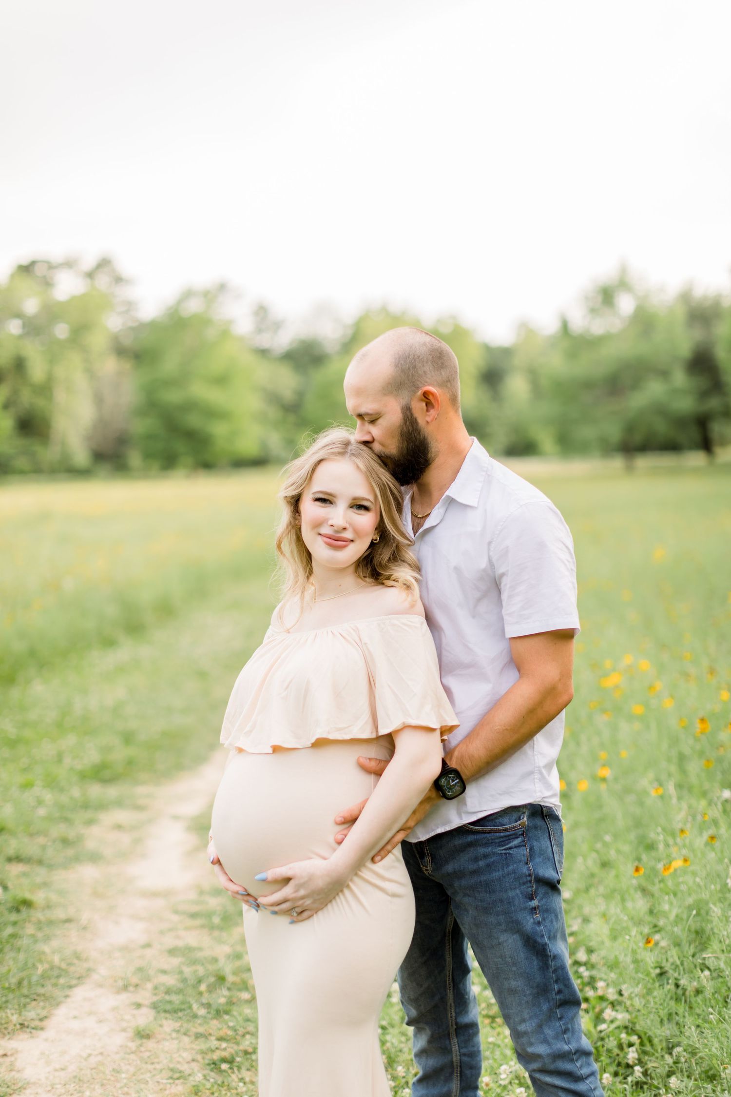 A maternity photo shoot in a sunny field with yellow wildflowers features an expectant couple in casual attire.