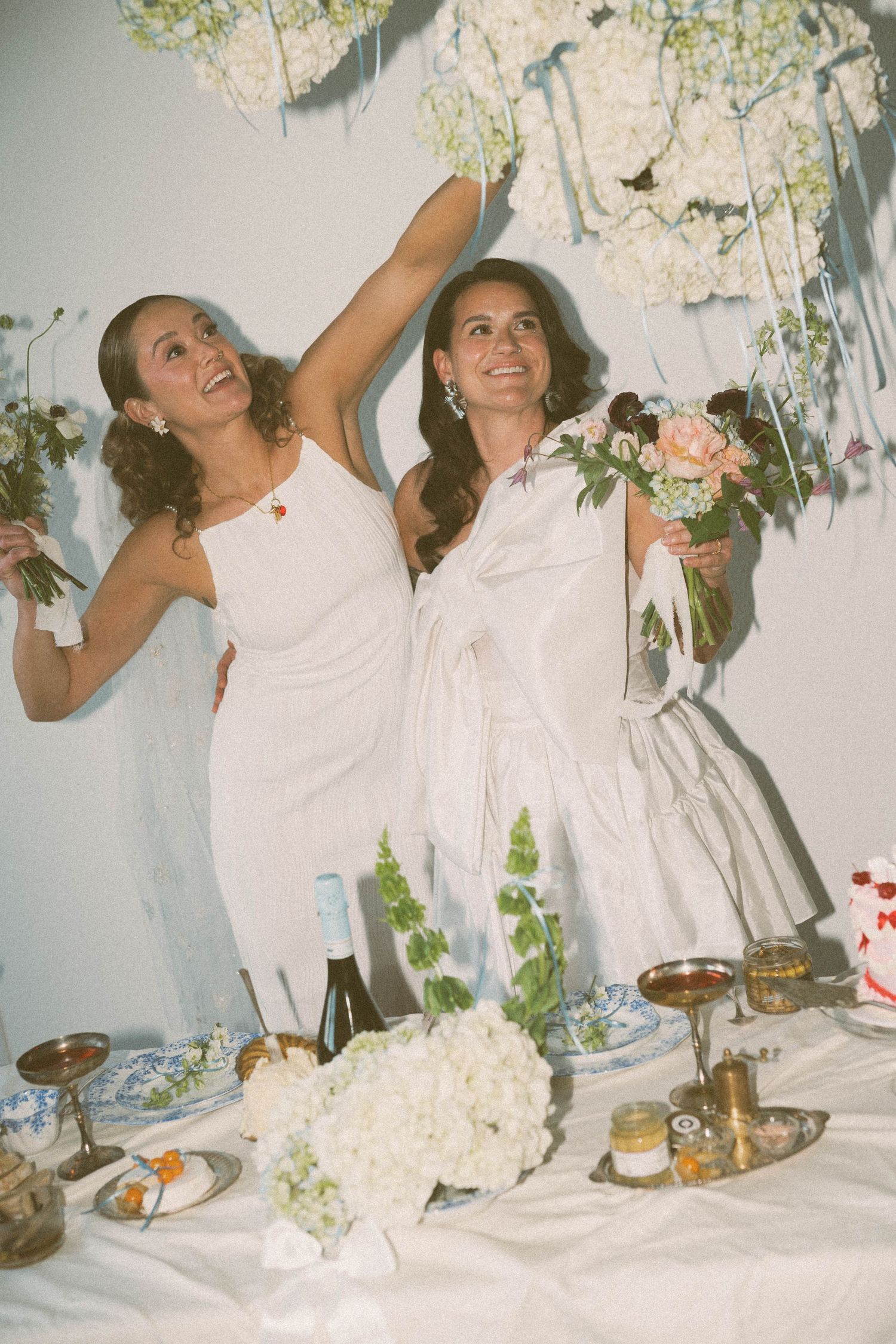 Two women in white dresses celebrating at decorated table with flowers and treats.