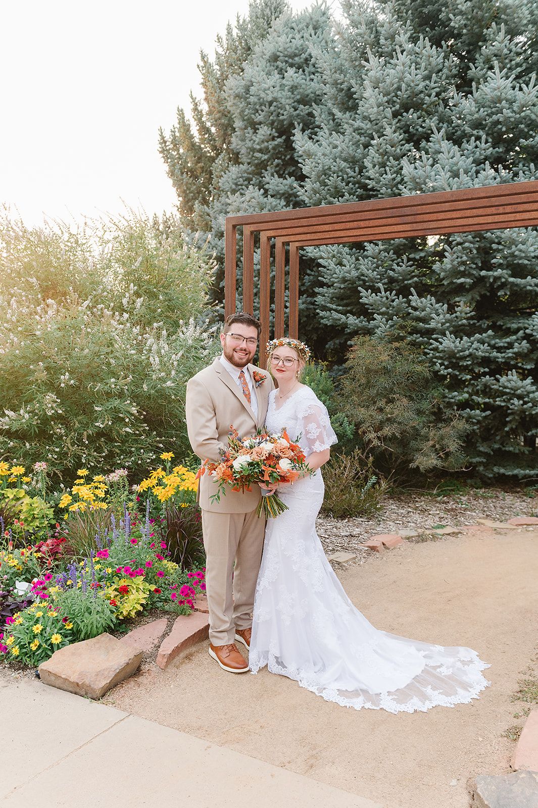 A wedding couple stands under a wooden pergola surrounded by colorful garden flowers.