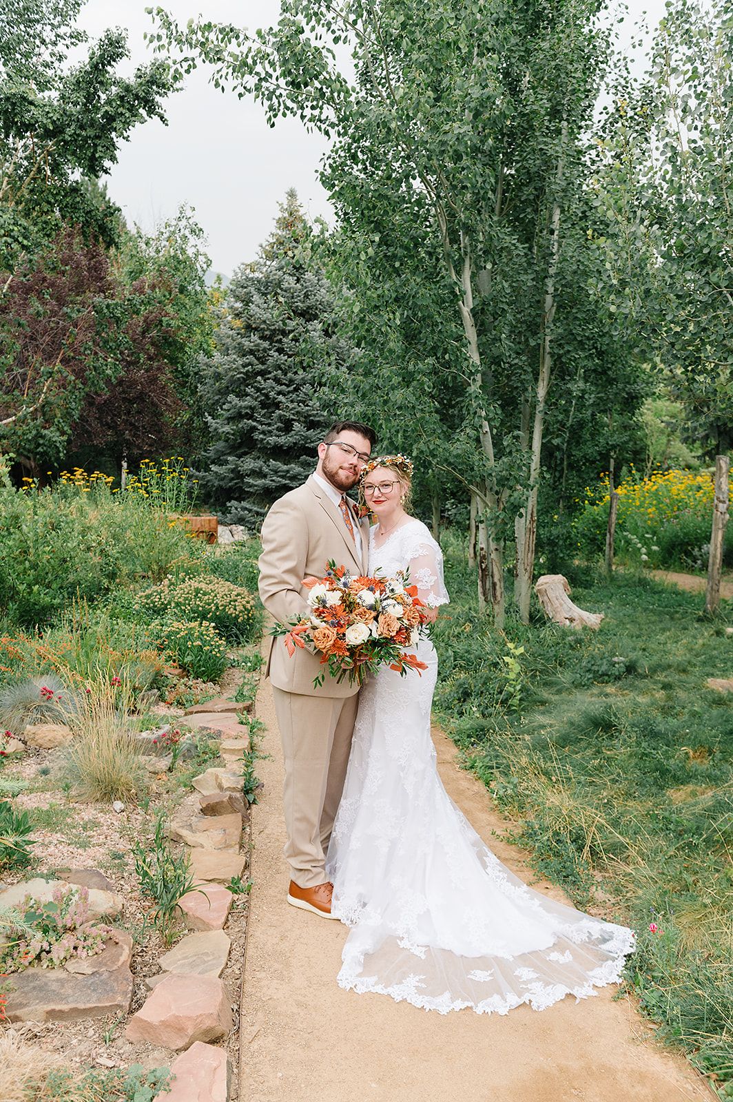A romantic wedding portrait in a lush garden setting with colorful flowers and tall trees.
