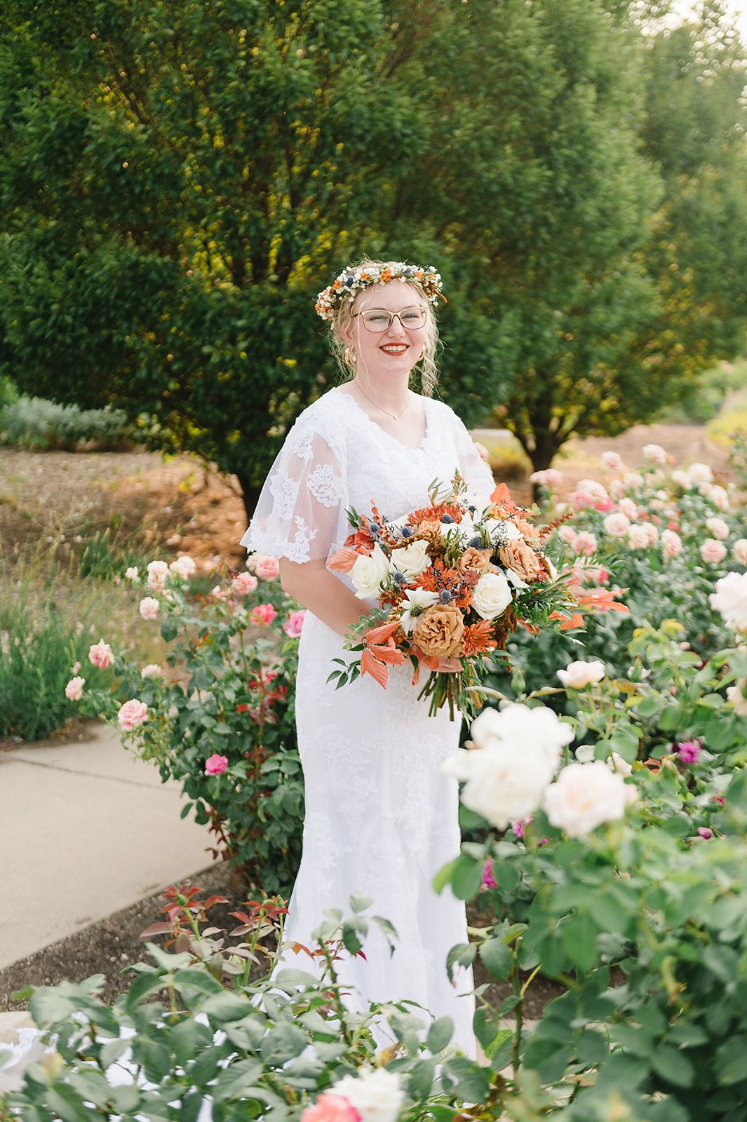 A bride in a white lace dress poses in a rose garden wearing a floral crown and holding a peach bouquet.