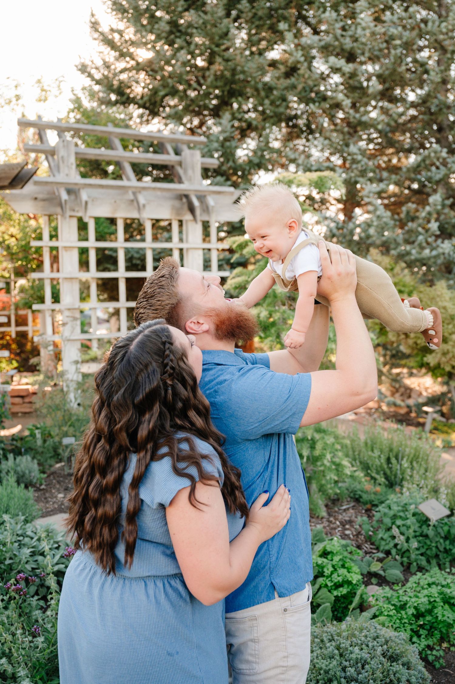 Parents lift and play with their infant in a garden setting with a white trellis arbor during golden hour.