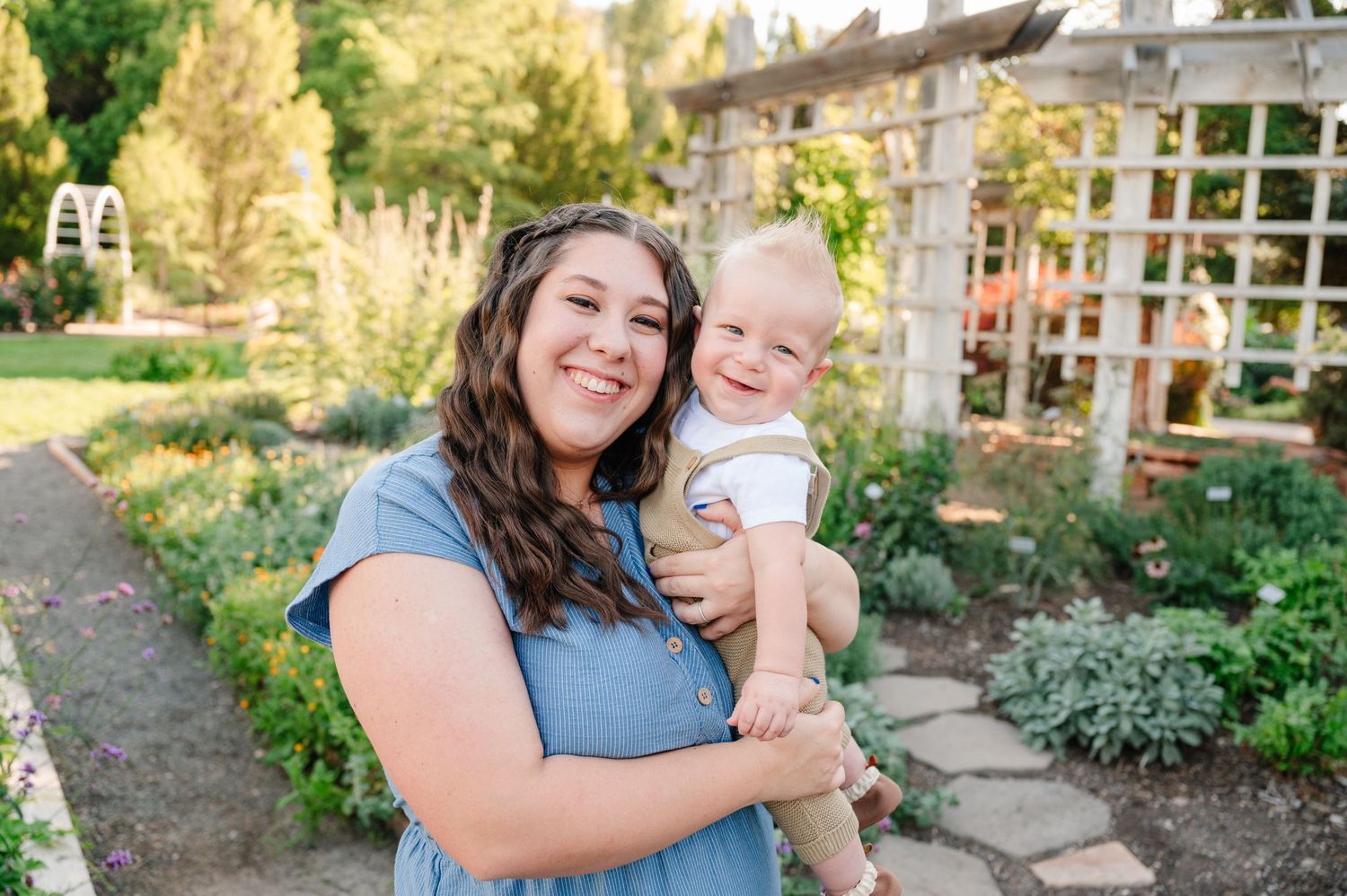A baby smiles while being held in a lush garden with a white trellis in the background.