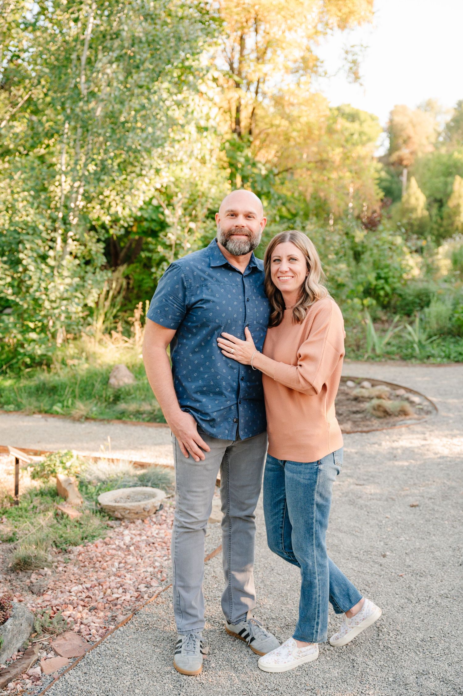A couple poses together outdoors in casual attire against a wooded background during sunset.