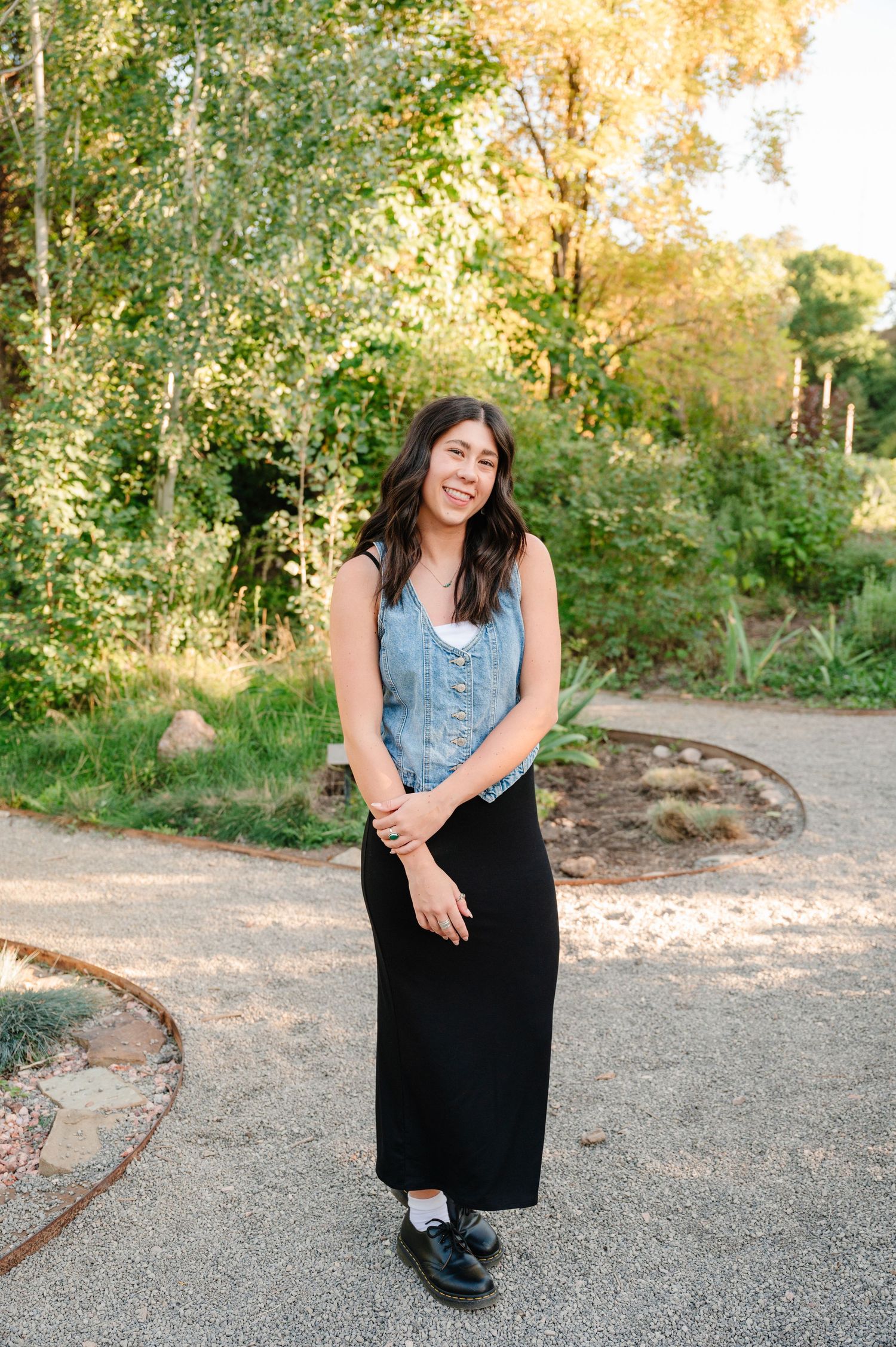 A woman in a denim vest and black maxi dress stands on a garden path during sunset.