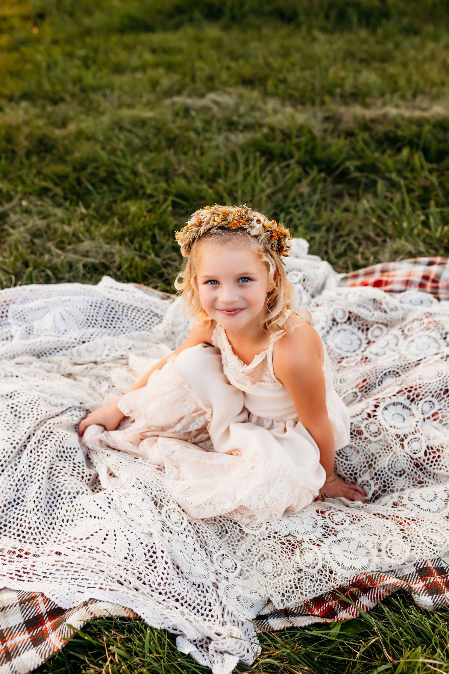 A child in a lace dress and flower crown poses on vintage blankets in a field.
