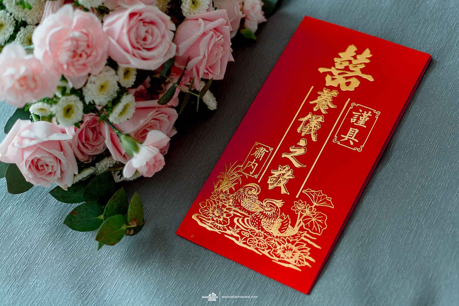 Traditional red Chinese wedding envelope displayed next to pink roses on blue surface.