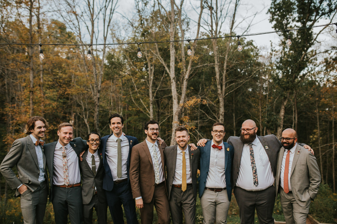 A group of formally dressed people standing together outdoors with autumn trees in the background.