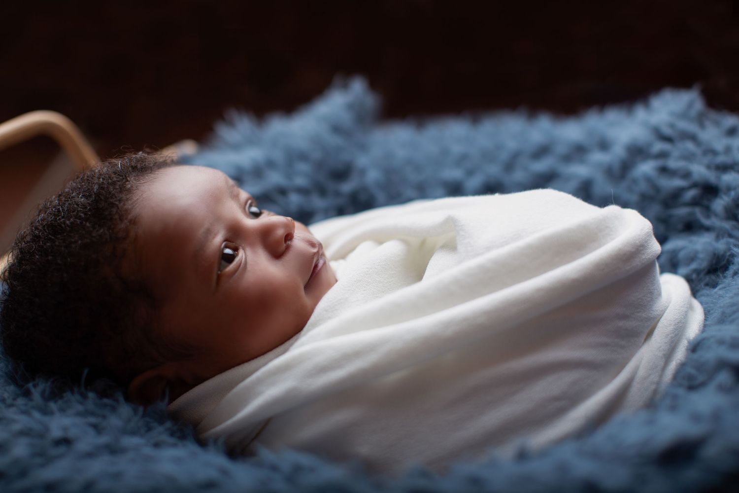Newborn baby swaddled in white blanket resting on blue fluffy surface.