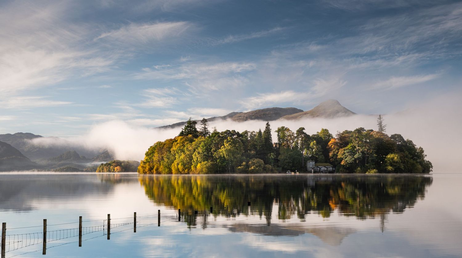 Misty lake landscape with tree-covered island reflecting perfectly in calm waters.