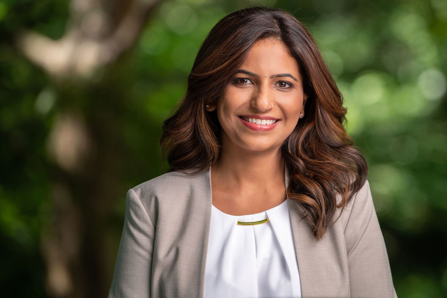 Professional headshot in white blouse and gray blazer against blurred green background.