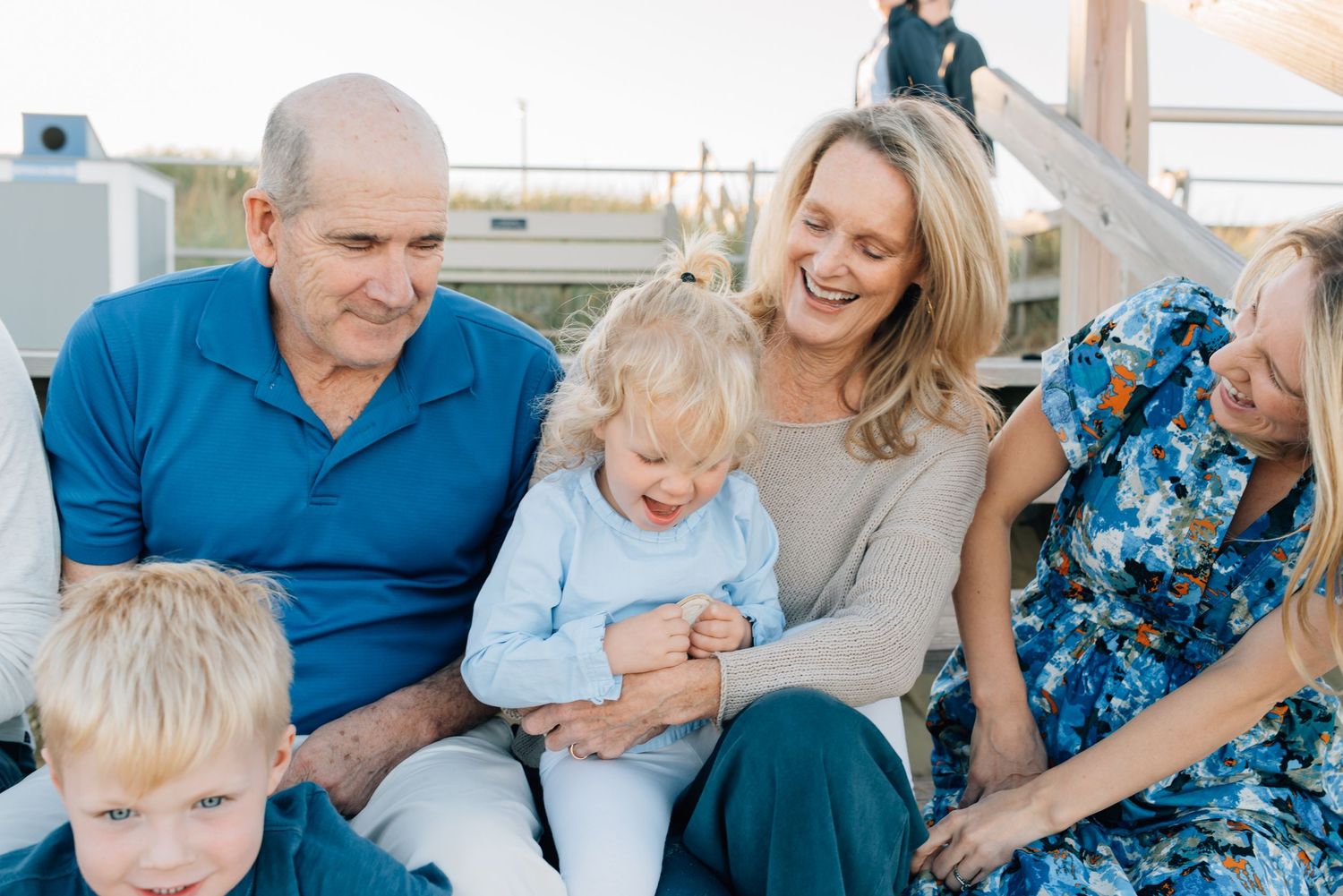 A grandmother and grandfather giggle with their grandchildren while sitting on the steps down to the beach.