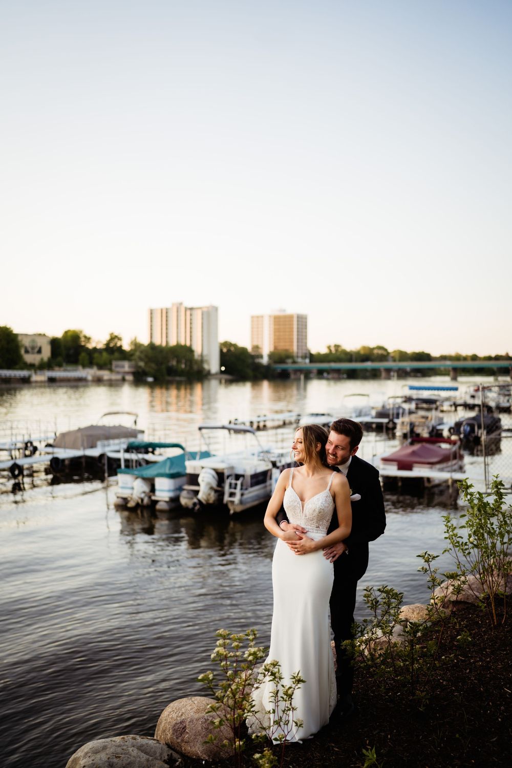 Couple embracing by waterfront with boats and city buildings in background at sunset.