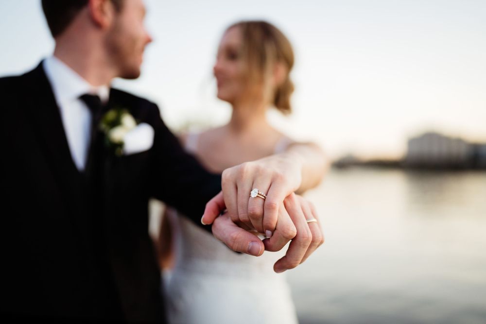Close-up shot of newlyweds' hands showing wedding ring against a sunset waterfront backdrop.