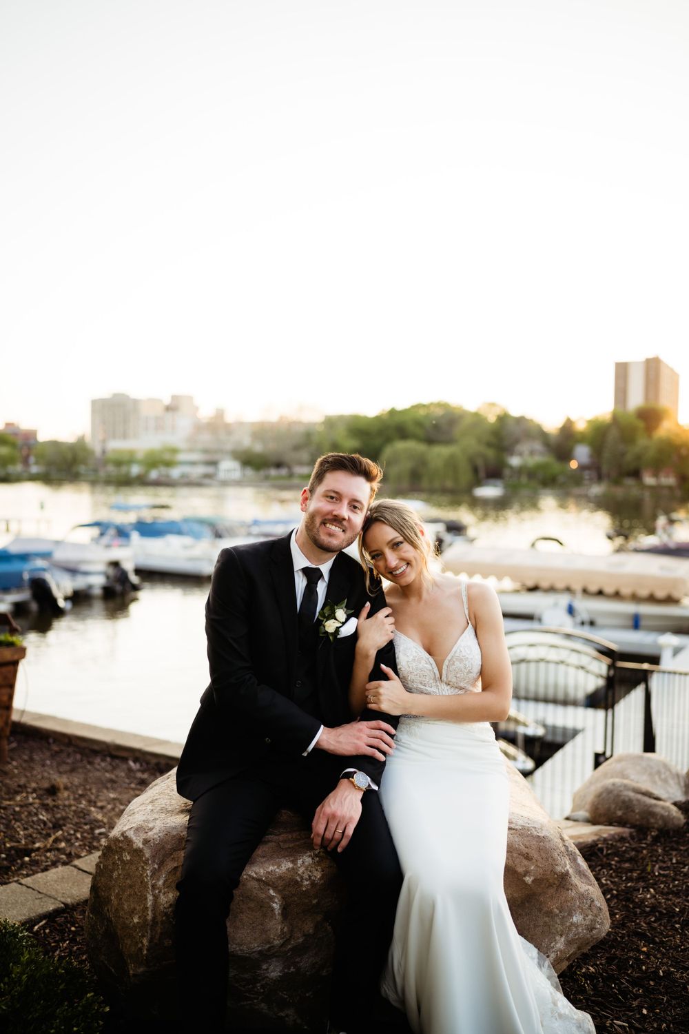 Couple sitting together on rocks by marina at sunset in formal attire.