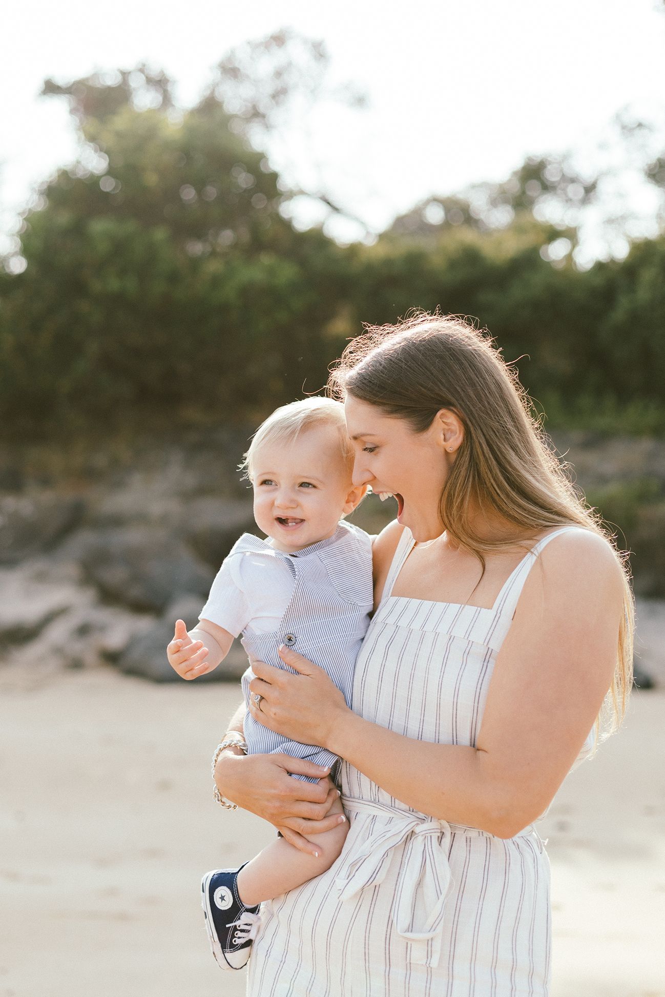 Mother in white sundress holds smiling baby during sunset beach photoshoot.