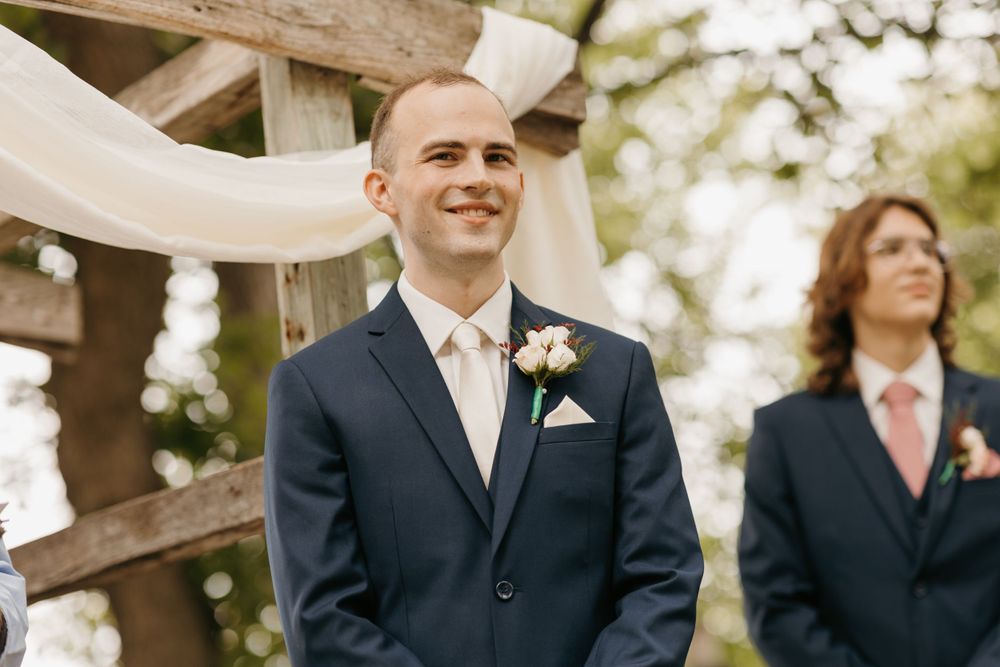 Groom smiles joyfully as bride approaches at rustic outdoor wedding ceremony altar.