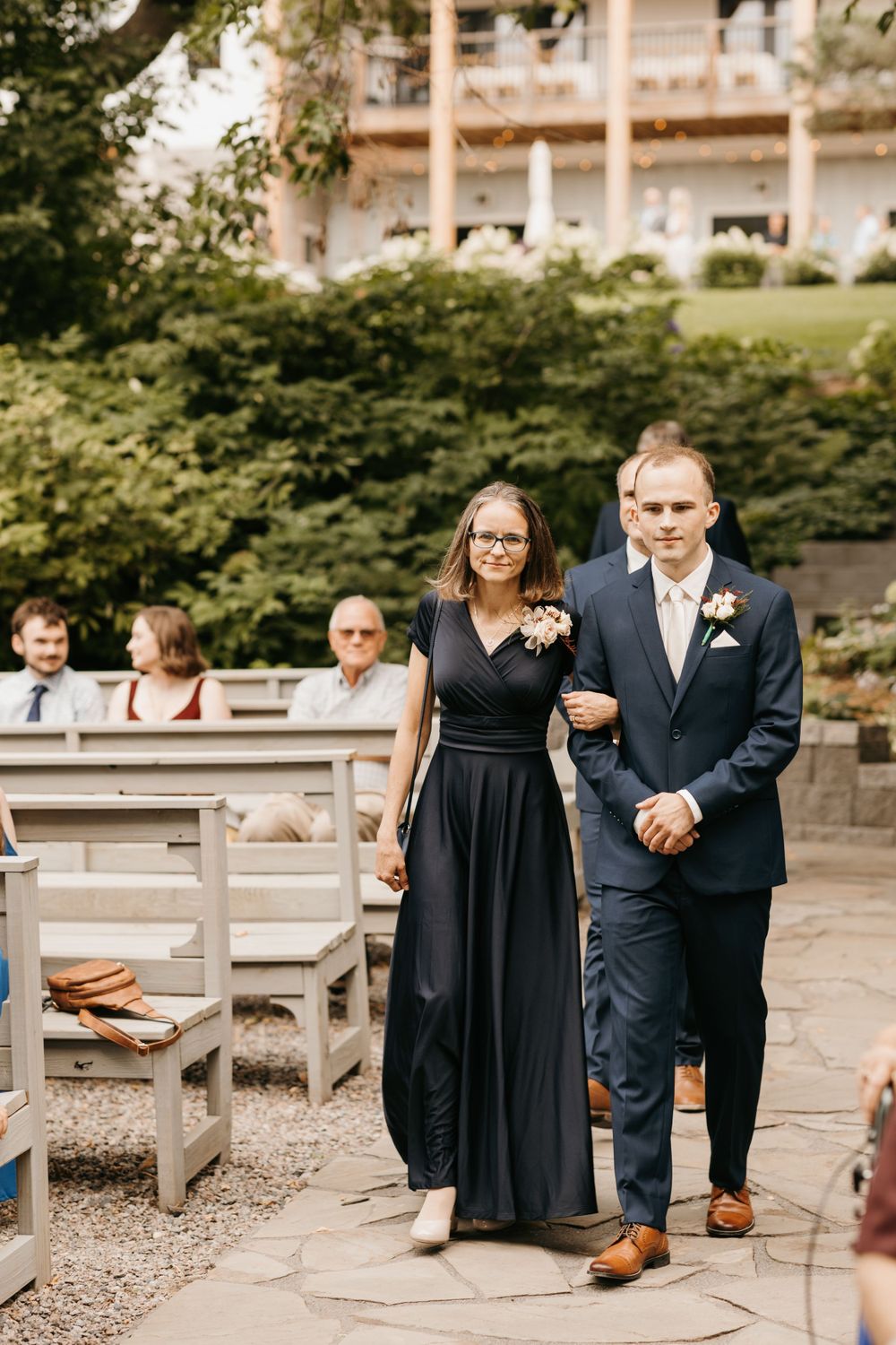 Groom escorts his mother in a black wrap dress down the aisle at an outdoor garden wedding.