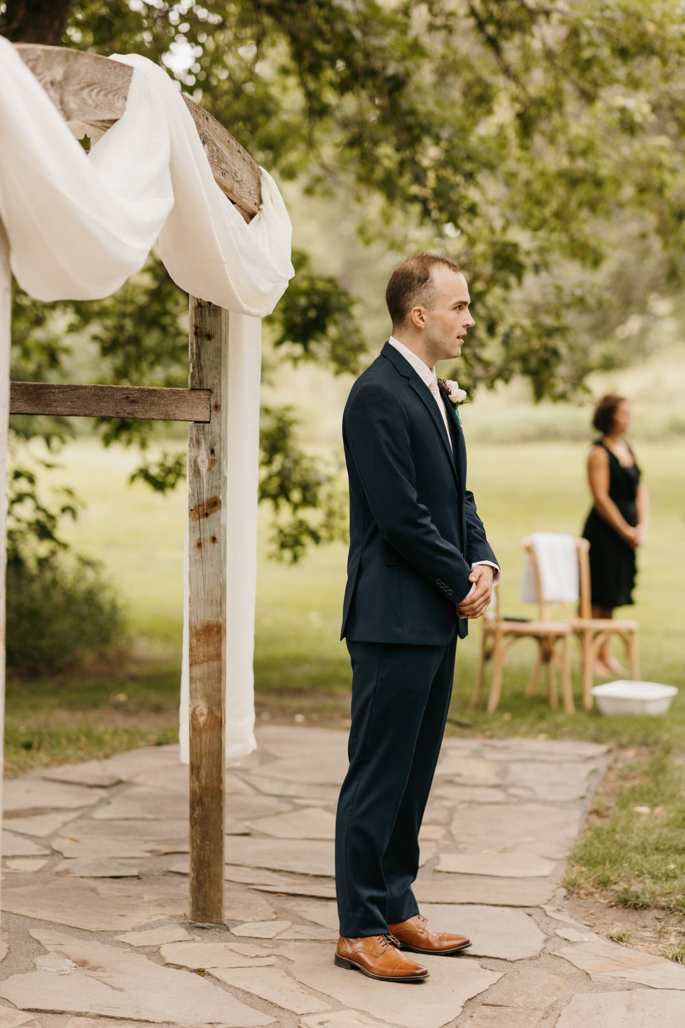Groom stands alone at rustic wooden altar with white draping at outdoor wedding ceremony.
