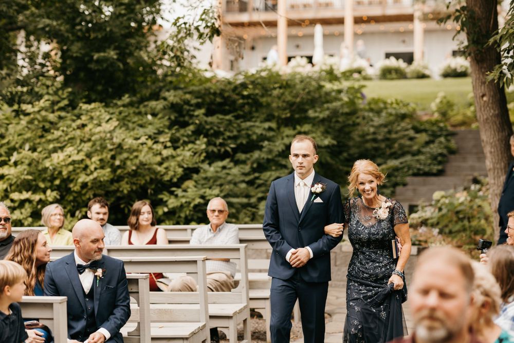 Groom walks down outdoor wedding aisle escorted by his mother in a navy sequin dress.