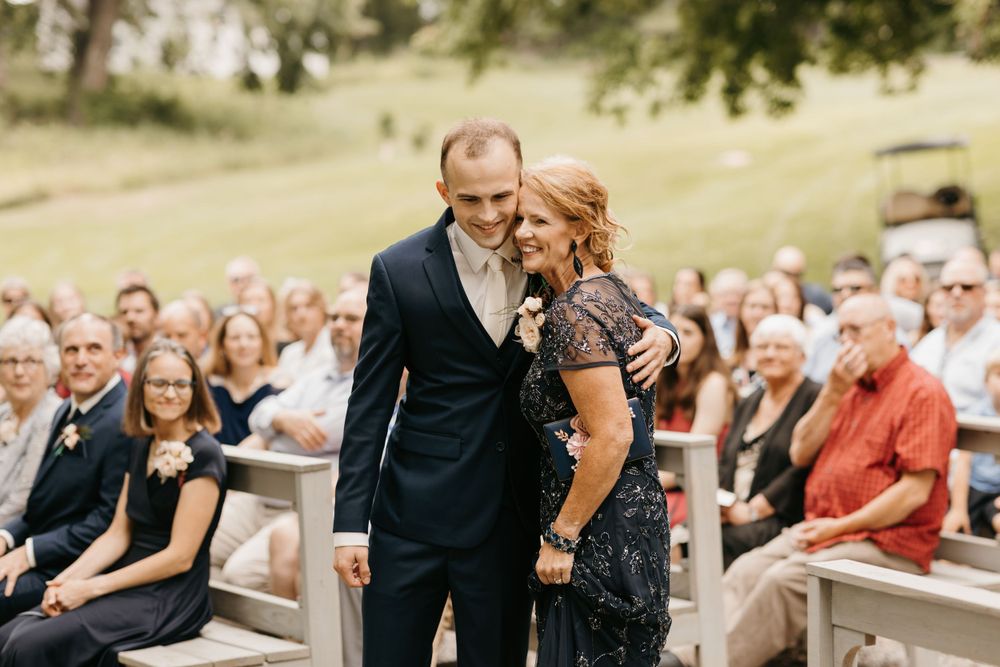 Groom smiles warmly while walking arm-in-arm with his mother at an outdoor wedding ceremony.