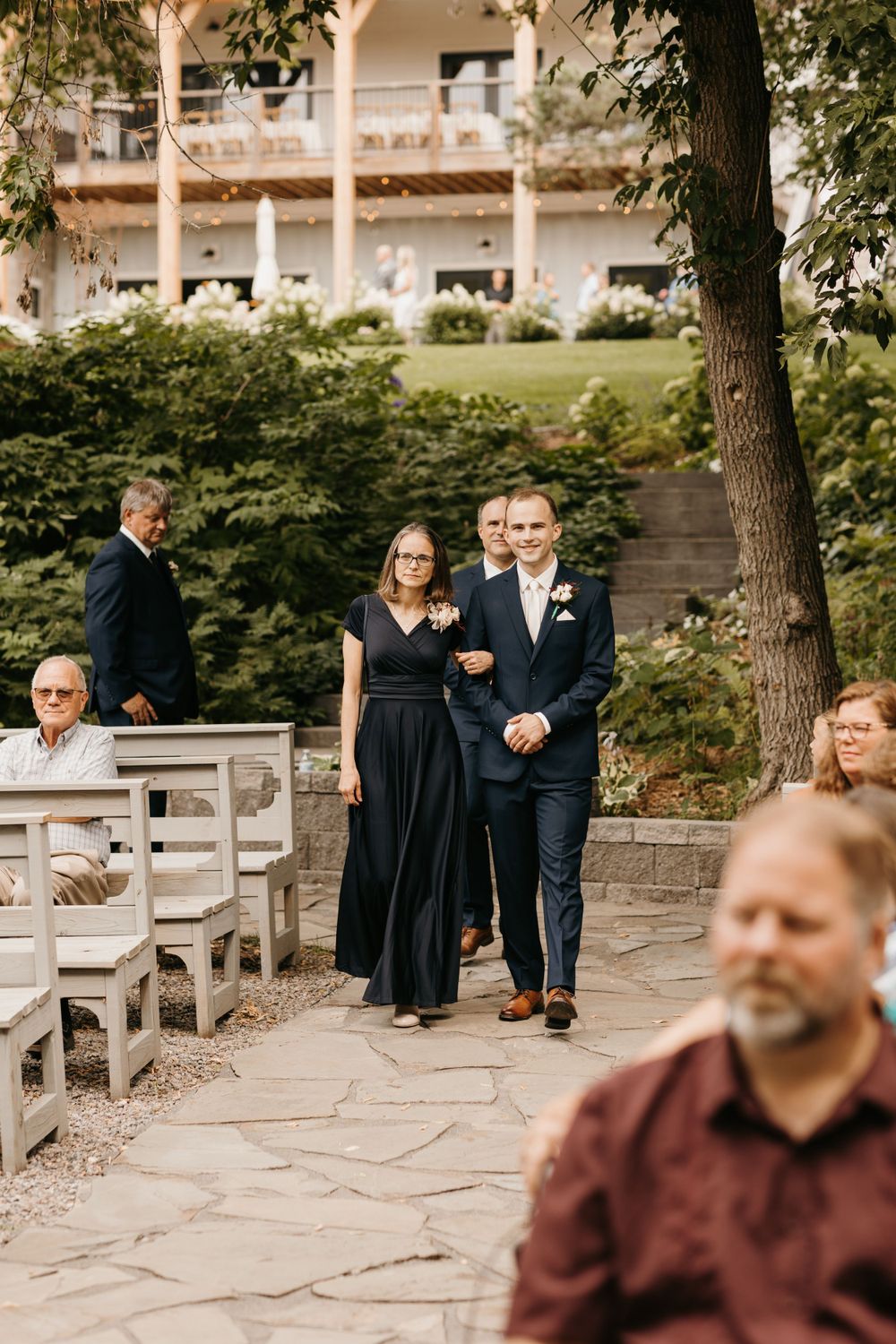 Groom walks down the aisle with his mother in a black gown at an outdoor stone wedding ceremony.