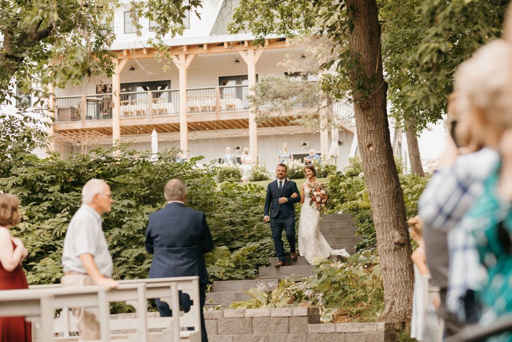Bride and escort walk down stone pathway toward wedding altar surrounded by lush greenery.