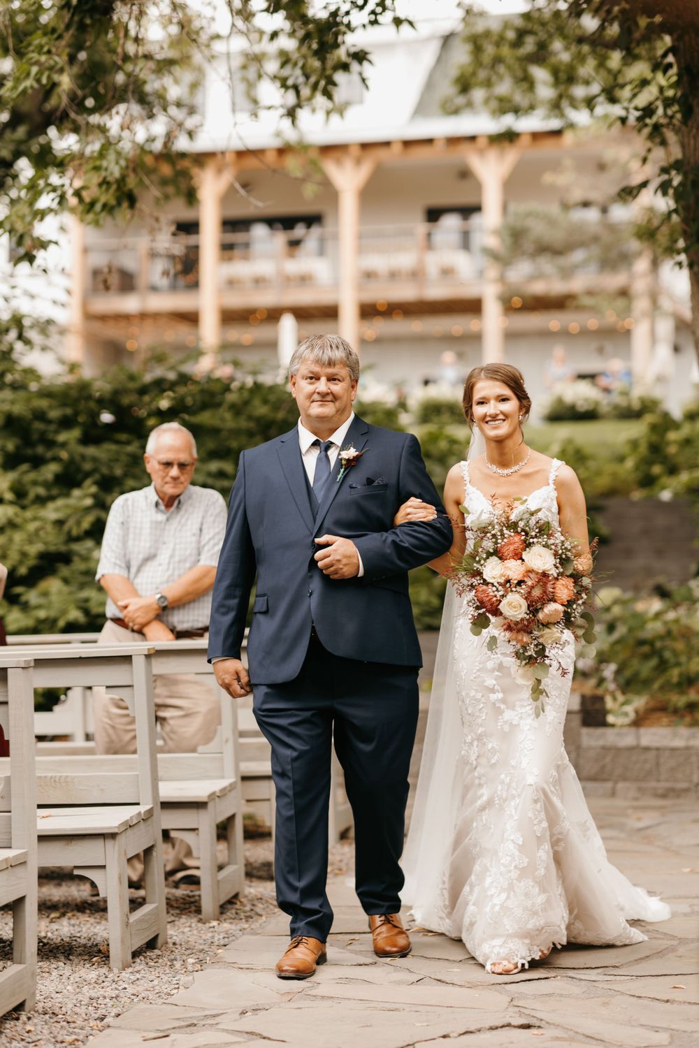 Smiling bride holding floral bouquet walks down aisle with father at outdoor wedding ceremony.