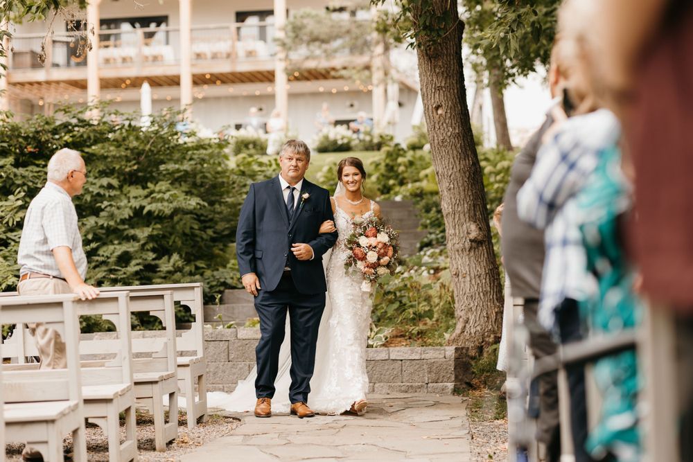 Bride in white gown walks down aisle with escort at elegant outdoor garden wedding ceremony.
