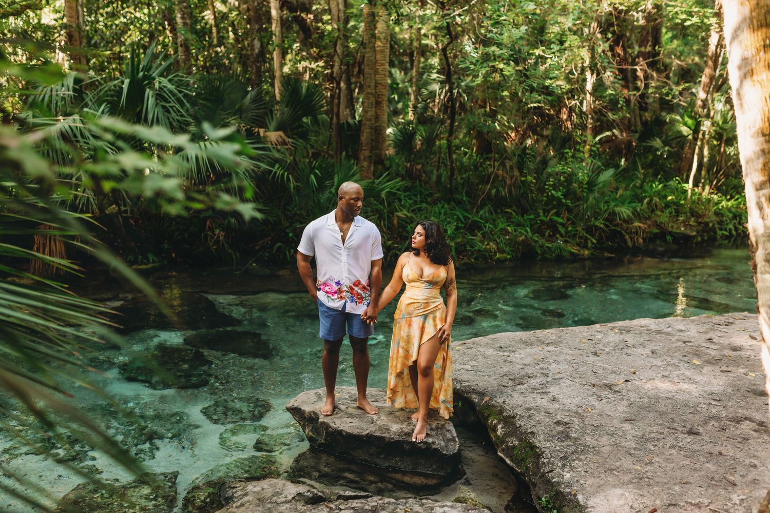 A couple stands by a crystal clear turquoise spring surrounded by lush tropical vegetation.