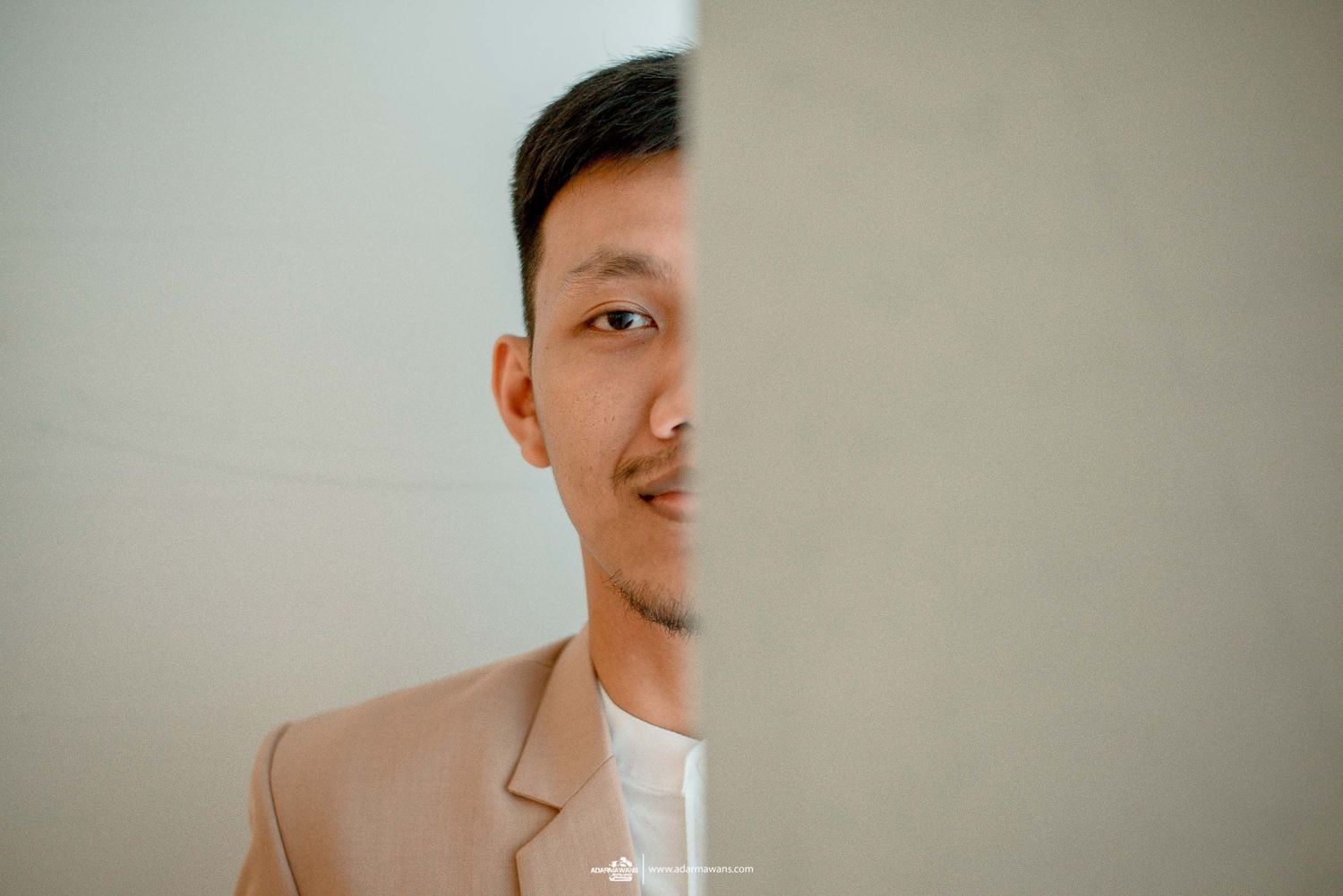 Creative portrait of man in beige blazer peeking from behind white wall.