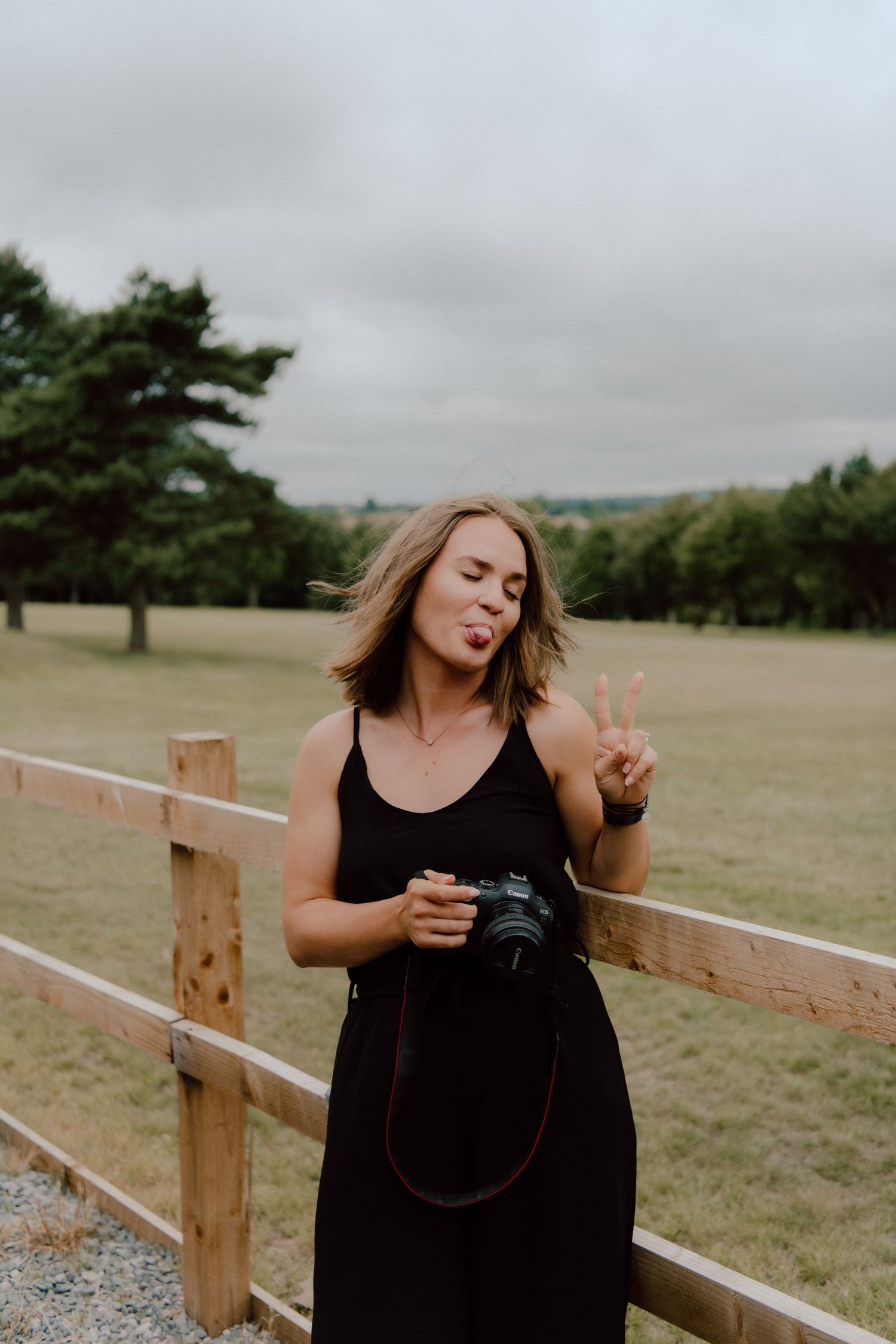 A photographer making a peace sign while holding a camera outdoors by a wooden fence.