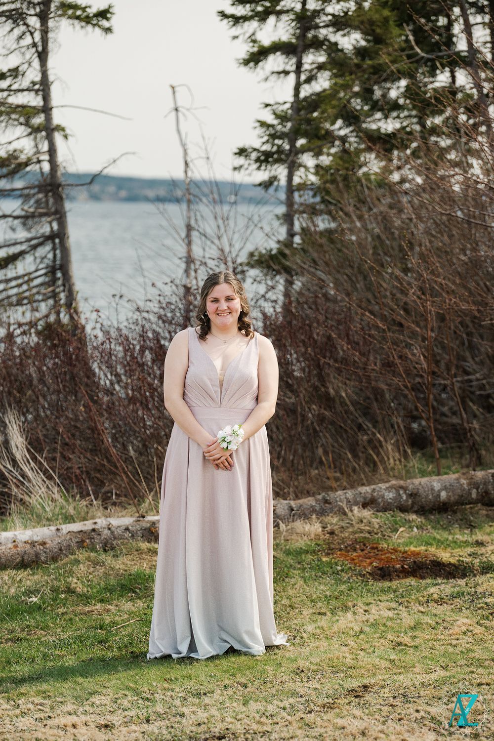 Woman in pale formal dress holding bouquet poses by waterfront with evergreen trees.