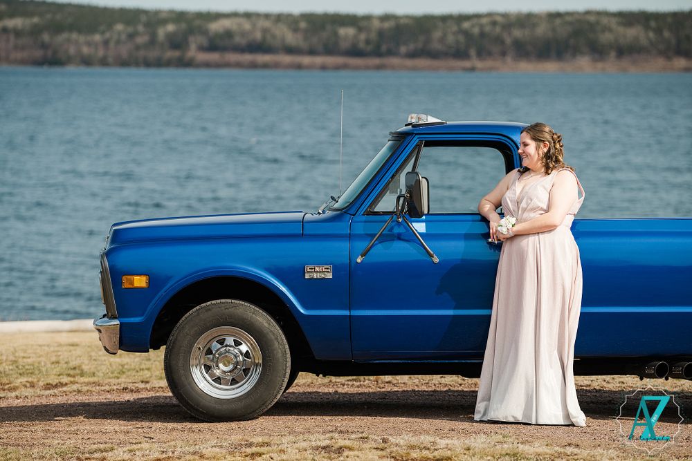 A white formal dress worn beside a classic blue pickup truck parked near a scenic lake.
