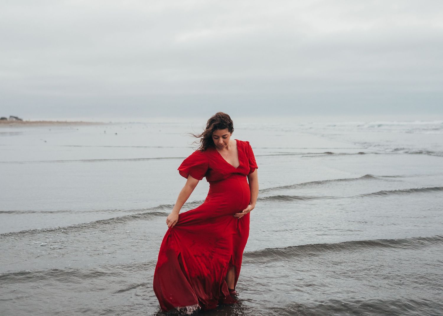 A woman in a flowing red dress walks along the shoreline on an overcast beach day.