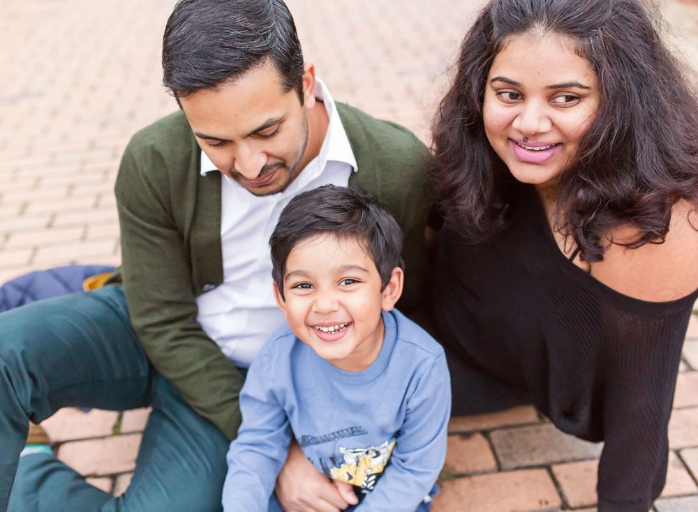 Candid family portrait with three people sitting on brick patio and sharing genuine smiles.