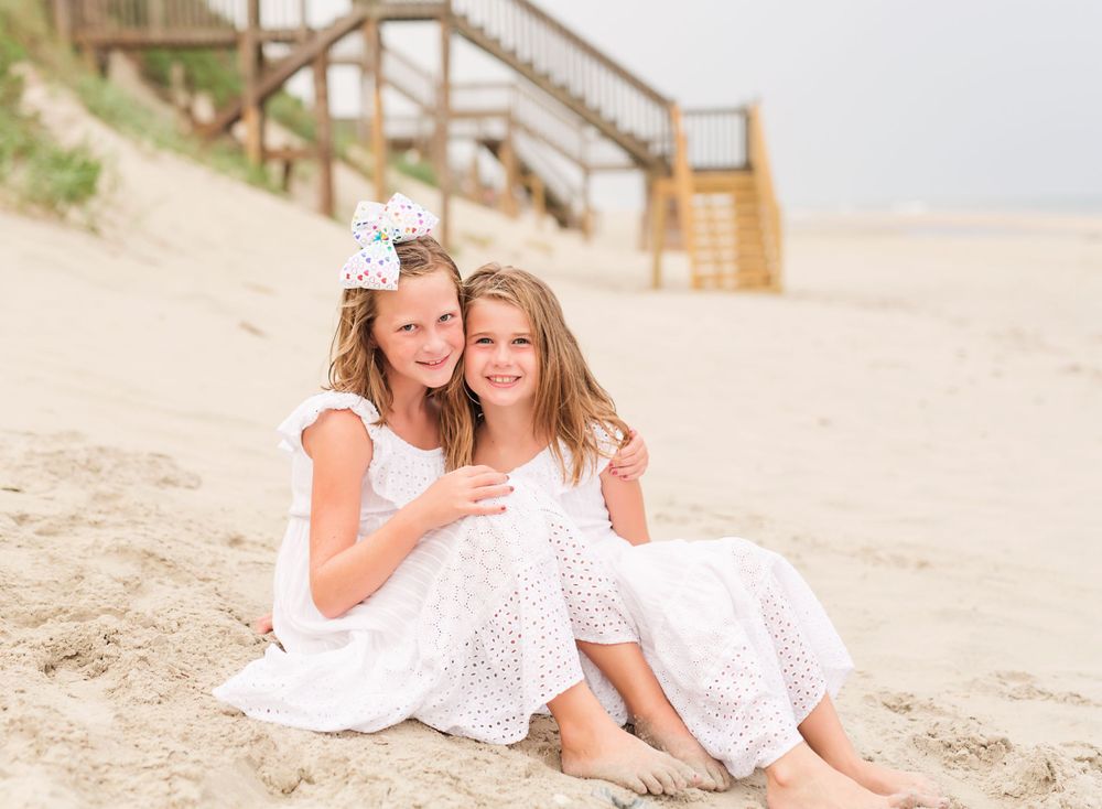 Two people in white dresses sitting together on sandy beach near wooden stairs.