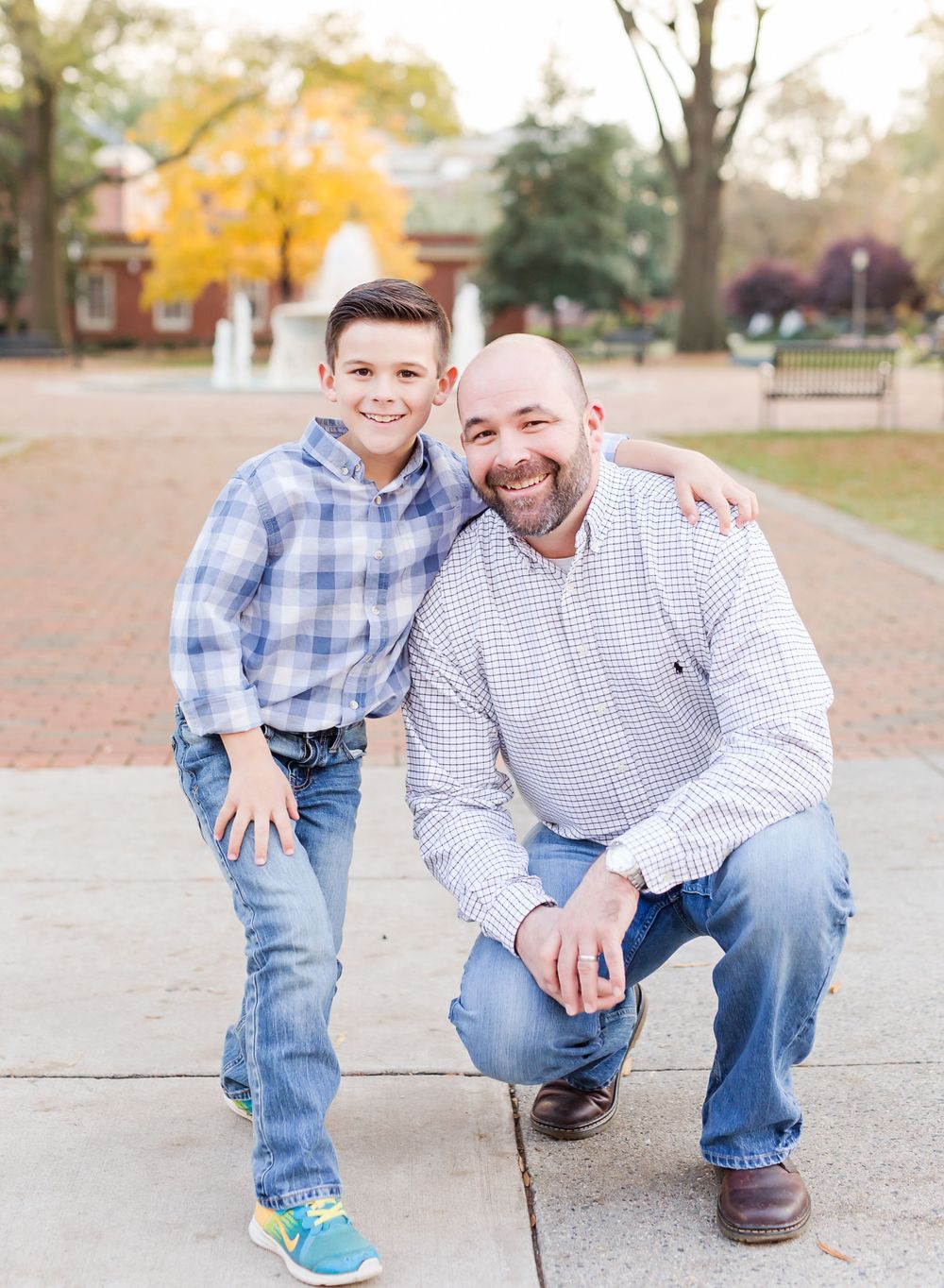 A father and son pose together on brick sidewalk with fall foliage behind them.